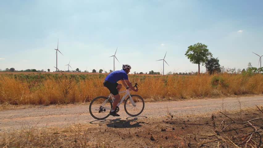 Drone view of active sportsman riding bicycle at the wind turbine field. Attractive male athlete in sportswear exercise by cycling workout for health and wellness in rural area at windmill power farm.