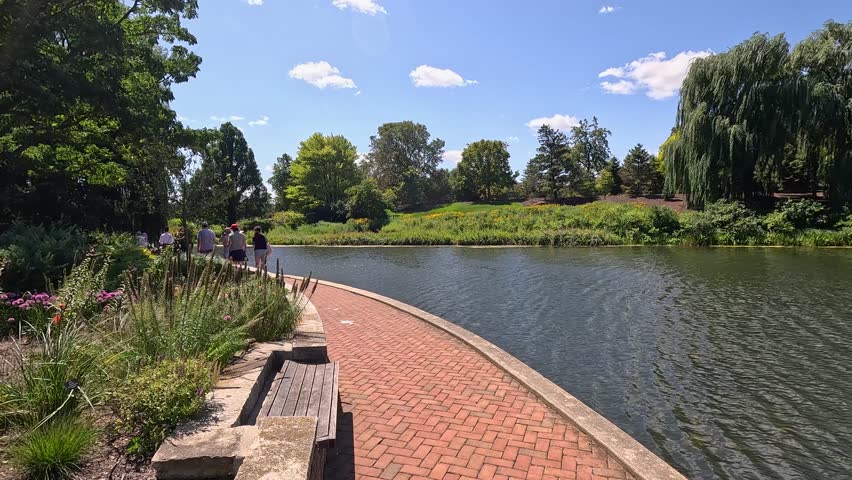 panning footage of a rippling lake with lush green trees, plants and grass, blue sky, clouds and people at Chicago Botanic Garden in Glencoe Illinois USA