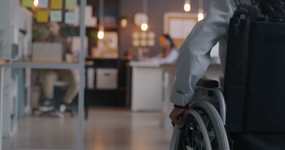 Back view slow motion of disabled businesswoman in wheelchair approaching workplace in office. Handicapped businessperson and inclusive workspace concept.