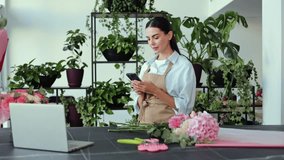 Cheerful young female florist stands at a counter in her flower shop, surrounded by colorful bouquets and lush green plants. Girl checks smartphone, managing orders or engaging with customers online. - Powered by Shutterstock - Get 15% off with code: PIKWIZARD15