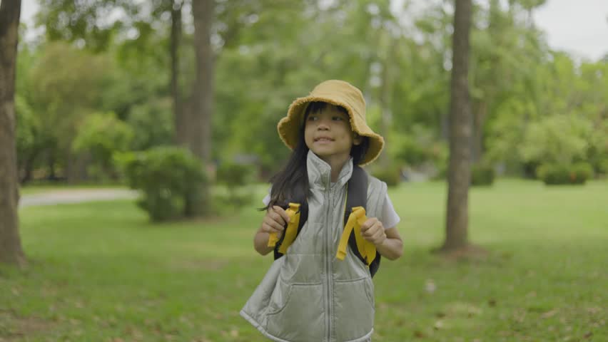 A cute little girl carries a backpack and walks around in a fresh green forest in different poses. She smiles happily as she goes out to study nature on her family vacation. Green forest background