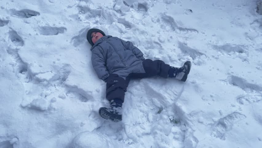 happy boy male child in gray winter jumpsuit lying snow making angel figure on white snow. baby waving hands and legs doing print of body angel figurine. happy christmas holidays outdoors games