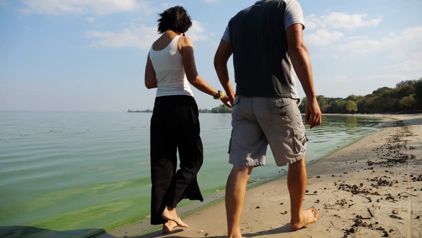 Couple holding hands of each other and walking at coast. Happy pair going along seashore at sunny day. Lovers spending time together at resort. Concept of vacation or holiday. Slow motion
