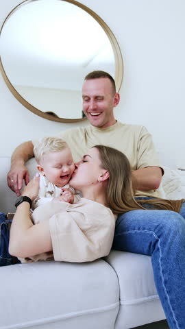 Happy Caucasian family at home. Man sits at sofa, his wife lies on his knees kissing her lovely plump baby boy. Vertical video.
