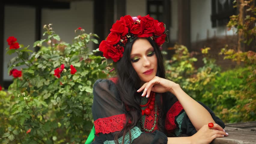 portrait happy girl sitting near house yard enjoys rest. authentic woman slavic beauty ukrainian traditional black dress flower wreath red beads, pretty face long dark hair festive makeup eyes lips. 