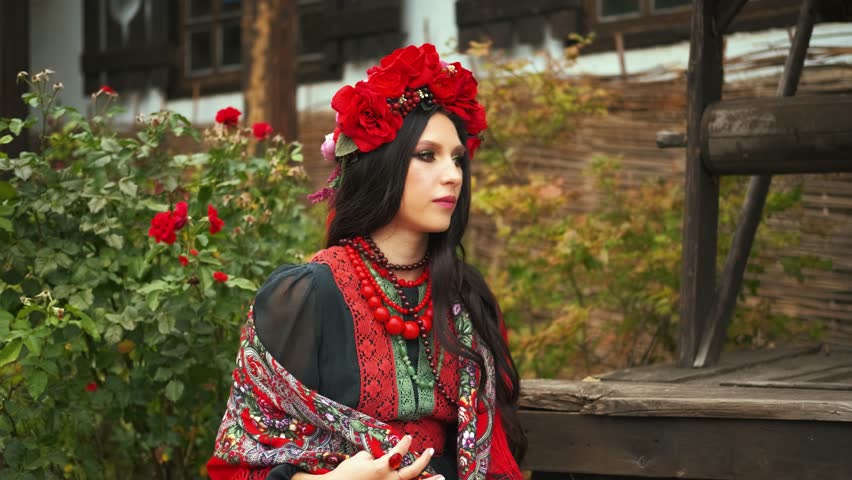 portrait happy girl sitting near house well yard. Young woman slavic beauty ukrainian black dress wreath red beads, pretty face long dark hair. Lady wraps herself in flower warm scarf cold autumn wind