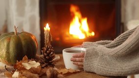 Autumn hygge. Hands in sweater holding cup of warm tea and pumpkins, autumn leaves on table against burning fireplace in living room. Cozy fall footage. Thanksgiving and Halloween. - Powered by Shutterstock - Get 15% off with code: PIKWIZARD15