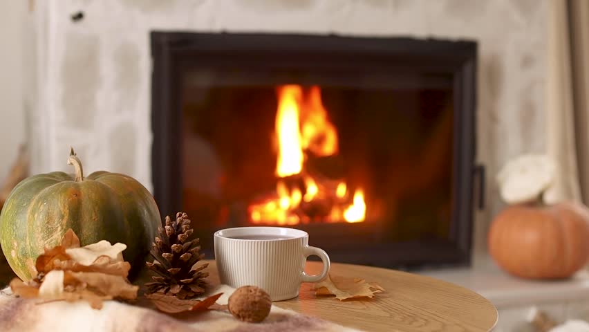 Autumn hygge. Cup of warm tea, pumpkins, autumn leaves on wooden table against burning fireplace in living room. Cozy fall footage. Thanksgiving and Halloween.