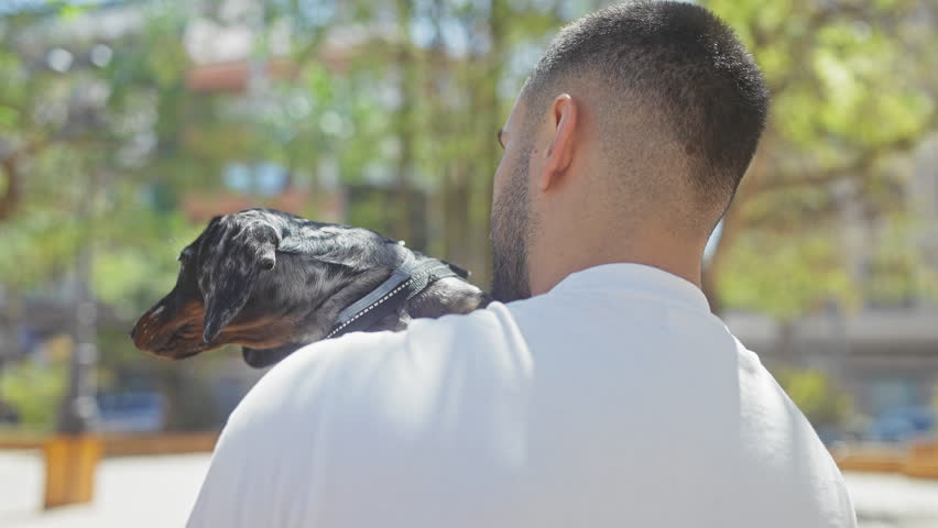 A young hispanic man in a casual white shirt lovingly holds his black and tan teckel dog in an urban park setting.