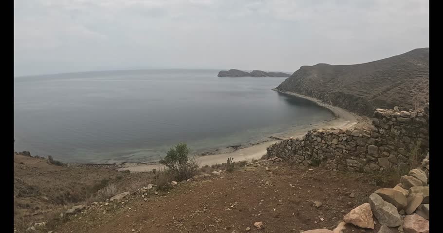 From the top of a hill, the view of Lake Titicaca is stunning. The blue waters stretch to the horizon, reflecting the clear sky. The lake shore is dotted with small boats and vegetation.