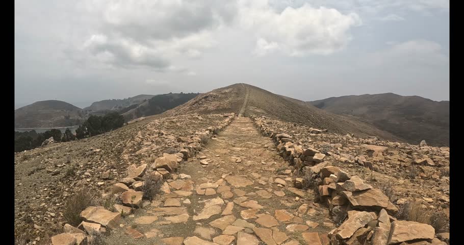 A video shows a stone path in Copacabana, Bolivia, winding along the shores of Lake Titicaca. The scenery is stunning, with mountains in the background and the tranquility of the blue waters. 
