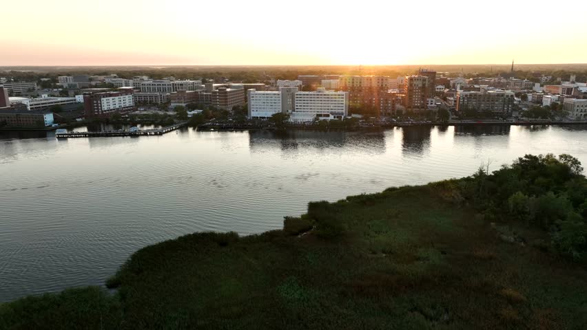 Aerial view of Wilmington, NC at sunrise, showcasing the city skyline and buildings along the Cape Fear River with the sun illuminating the horizon.