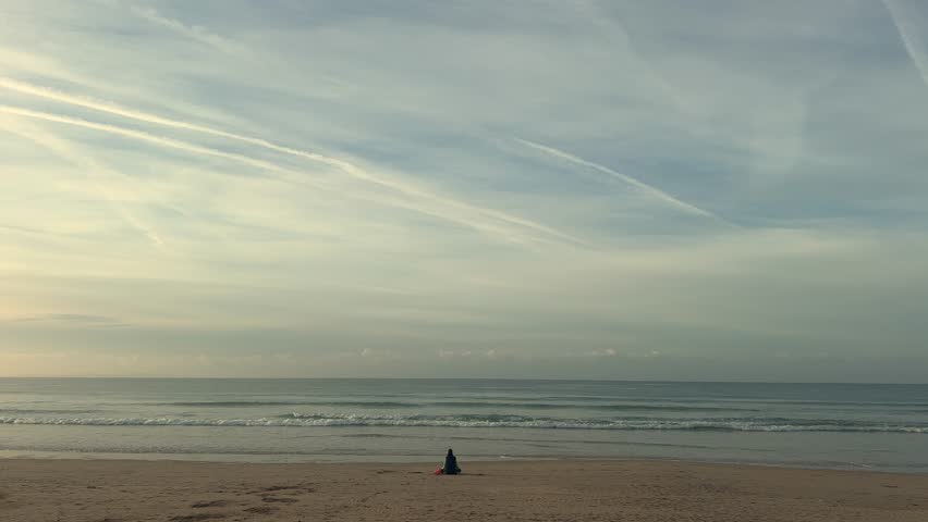 A lone person sits on the beach, gazing at the distant sea horizon. The scene, set in coastal Spain, embodies peace and serenity, capturing a sense of solitude and calm reflection.