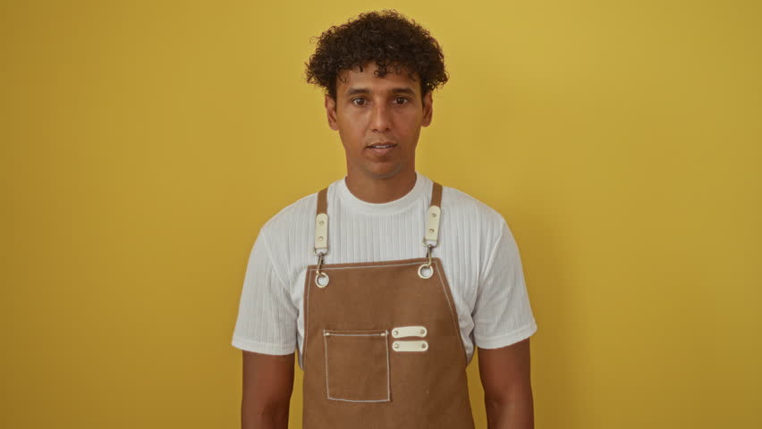 Young hispanic man in brown apron over isolated yellow background gesturing expressively