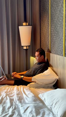 asian man working with his computer in bed, happy and smiling after receiving a message on his cell phone.