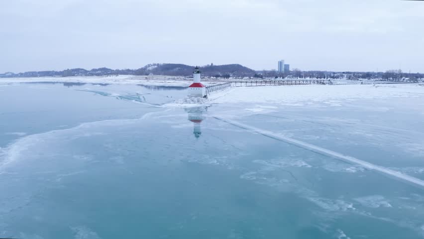 Aerial views of Frozen harbor and Lake Michigan and Michigan City red lighthouse and pier. Smooth and choppy ice surrounding the pier and coastline.