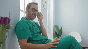 A mature hispanic male doctor listens to a voice message on his phone in a hospital waiting room, dressed in green scrubs with a stethoscope around his neck, during a quiet moment inside. - Powered by Shutterstock - Get 15% off with code: PIKWIZARD15