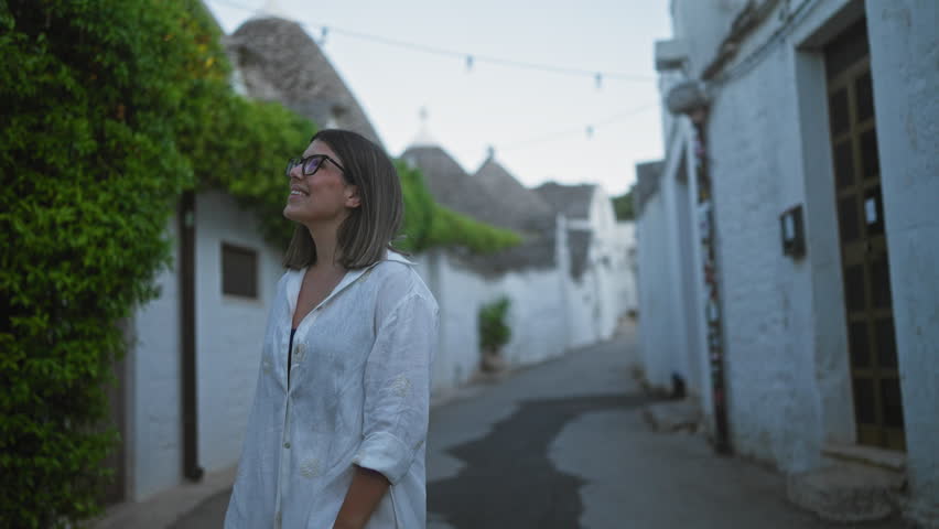 A young hispanic woman in a white shirt smiles as she walks down a charming street in the old town of alberobello, italy, surrounded by the traditional trulli houses.