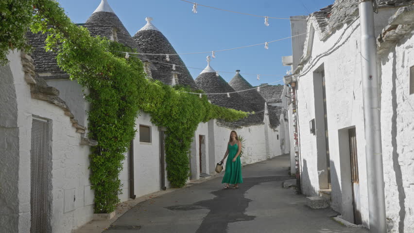 Young hispanic woman in a green dress walking in the charming old town of alberobello, italy, surrounded by iconic trulli houses and vibrant greenery under a clear blue sky.