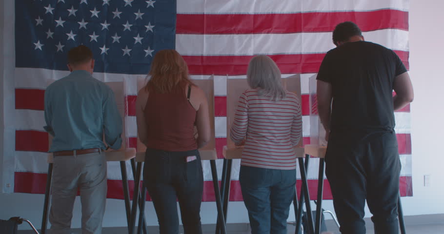 Wide shot of an  ethnically diverse group of voters voting at a polling station in front of a large American flag
