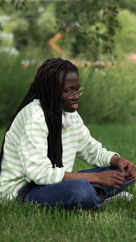 African American woman with dreadlocks sits cross-legged on grass in park, enjoys peaceful rest in fresh air and enthusiastically talks about thoughtful topics. Interested conversation between people.