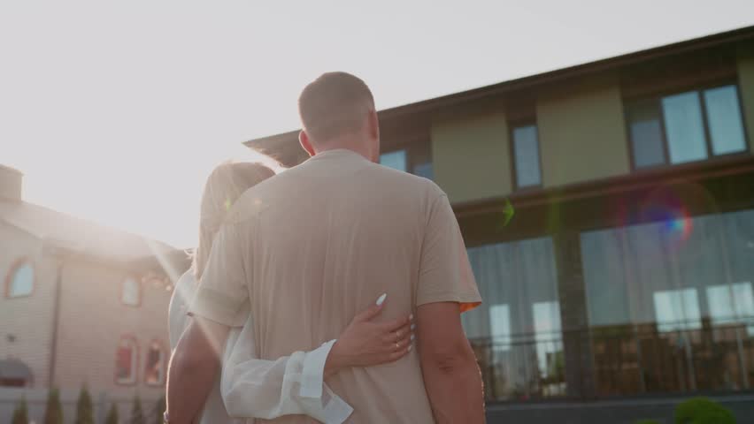 Back view of caucasian couple looking at new luxury house while standing at the yard. Young family dreaming about buying a new big house standing and hugging.