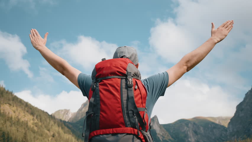 Back view gimbal shot man hiker with red backpack standing raised hands, male tourist raising up hands looking at beautiful breathtaking mountain landscape. Backpacker celebrate, hiking in mountains