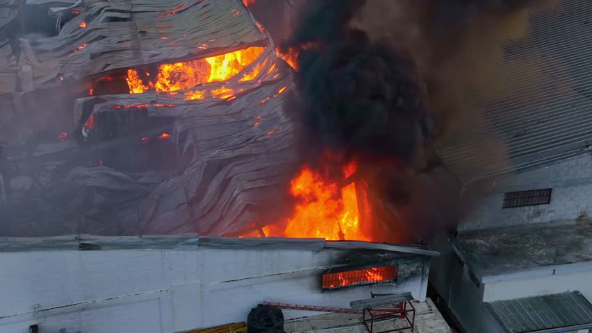 Zoomed-in aerial view of collapsed roof on active warehouse fire with dense black smoke