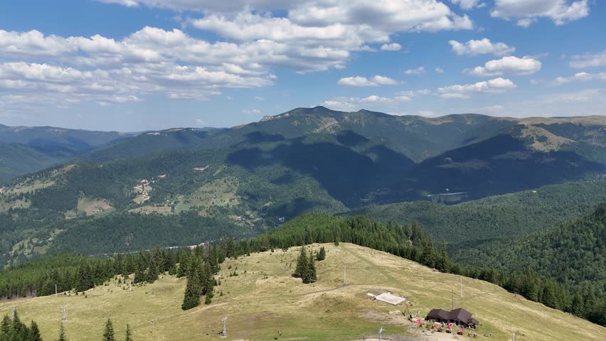 Rotating aerial view of Borsa’s landscape surrounded by tall mountain peaks, green fir forests, and a clear blue sky with white clouds, Bucovina region, Romania