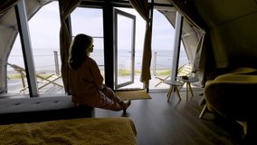A woman lounges comfortably on a bed, gazing out of large open windows that offer a stunning view of the beach and ocean. The serene setting blends the warmth of the cozy interior with the calming - Powered by Shutterstock - Get 15% off with code: PIKWIZARD15