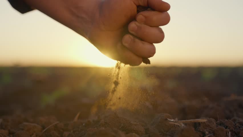 Closeup view of male hands with soil against beautiful agricultural field. Farmer in farmland, soil treatment and cultivation, agro industrial complex in eco region, timeless and endless, slow motion