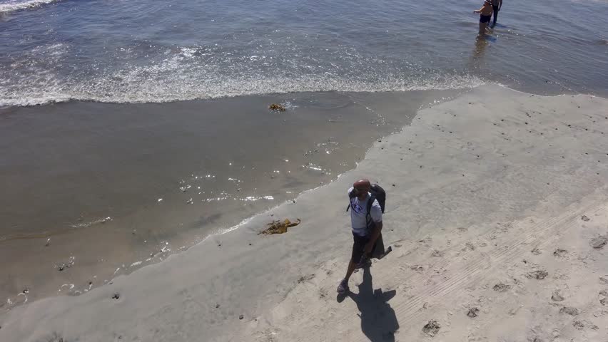 aerial footage of an African American man wearing a backpack walking along the silky brown sand at South Oceanside Beach with blue ocean water and waves and large rocks along the beach with blue sky