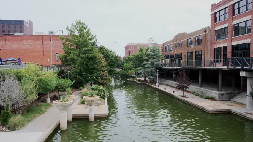Static wide shot of Bricktown canal with brick buildings, calm waters, and lush trees in Oklahoma City.