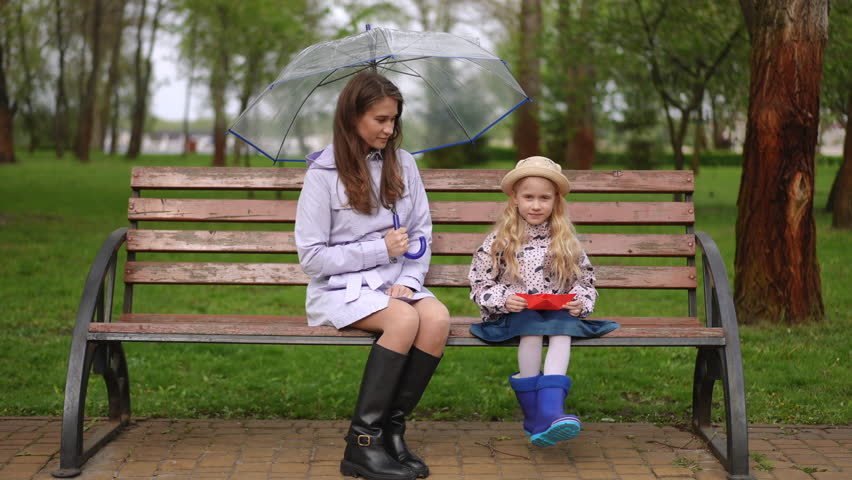 On this rainy day, a mother and her daughter share a peaceful moment on a cozy park bench under an umbrella