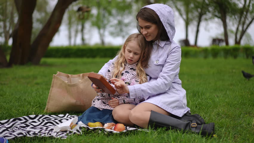A loving mother and her daughter are happily spending time reading together on a lovely picnic blanket in a serene park