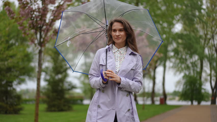 A graceful woman elegantly holding a transparent umbrella while enjoying a lush park during a light rain shower