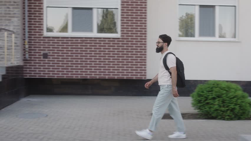 A young man wearing a white shirt, light pants, and glasses with a backpack walks up a set of stairs to enter a brick building.
