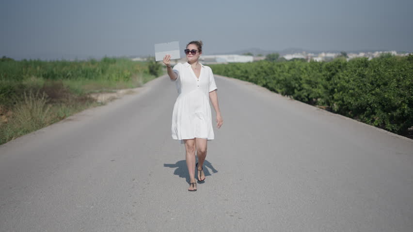 young curly haired blonde Caucasian woman in a short white dress walking facing the camera on a lonely suburban road gesturing while making a video call with her tablet