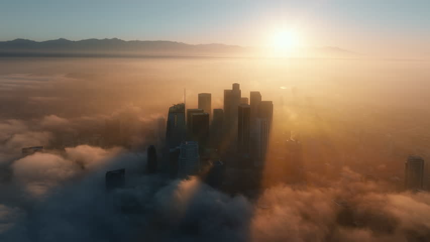 An aerial view of Downtown Los Angeles at daybreak reveals skyscrapers through morning mist, as the sun casts warm, golden light and enchanting shadows on the vibrant cityscape below