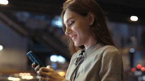 Beautiful Smiling Girl Walks Down the Night Street and Texts, Smiling at Her Phone. Positive Lovely Woman Uses Her Phone, Chats on Social Media. People and Technology Concept - Powered by Shutterstock - Get 15% off with code: PIKWIZARD15