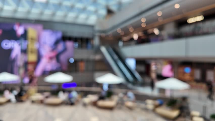 Blurred view of a modern shopping mall with an escalator, seating and large storefront displays.