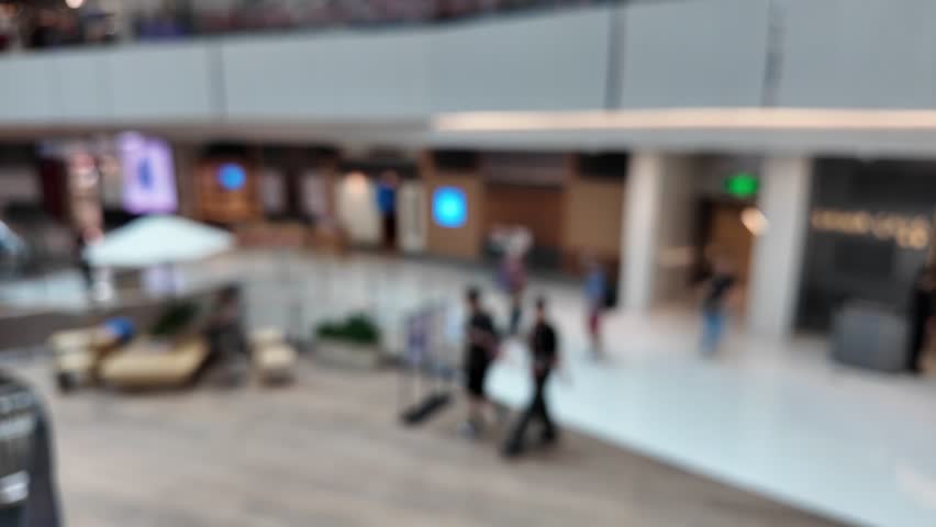 Blurry view of a shopping mall interior with people walking, stores, and a kiosk.
