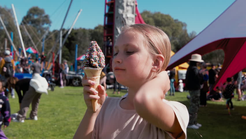 In a vibrant outdoor fair, a young girl savors a colorful ice cream cone, surrounded by families, attractions, and a festive atmosphere under the sun.