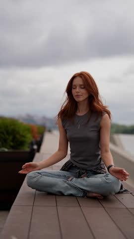 Vertical portrait of serene young woman with red hair meditating outdoors on city bridge, finding peace and mindfulness in urban environment. Relaxed redhead female finding inner peace and balance.