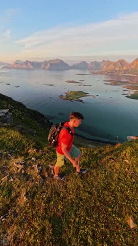 hiker with a backpack descends a grassy hill on the Lofoten Islands, Norway, during sunset. The vast ocean and distant islands are visible below, bathed in the golden light of the setting sun