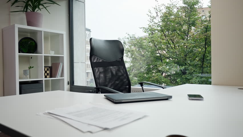 in modern office with a large window near a white table with a laptop there is a black office chair comfortable work flexible mesh back of the chair
