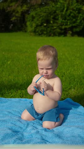 Vertical video of baby holding blue sunglasses and having fun in garden. Little boy playing on carpet on green grass in garden. Funny little child looking for something outside