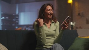 Excited beautiful Caucasian girl winning online prize in app. Cute young woman sitting on sofa and using smartphone. Female businesswoman celebrating victory in evening. Living room. Indoors - Powered by Shutterstock - Get 15% off with code: PIKWIZARD15