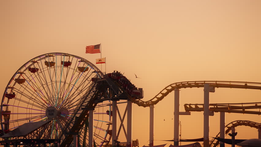 American flag waving and tourists riding a rollercoaster in Pacific Park Santa Monica pier in Los Angeles, California during sunset