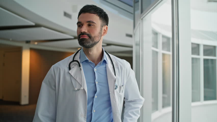 Confident doctor walking hallway with stethoscope. Closeup smiling man therapist looking camera going modern healthcare center in white coat. Young cheerful surgeon at work. Dedicated medical worker